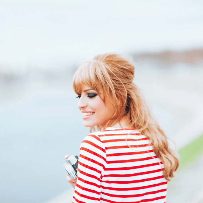 Vrouw met camera in een gestreept shirt aan de waterkant, glimlachend en zelfverzekerd.