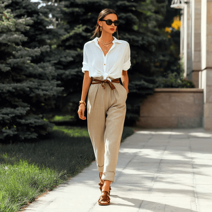 Stijlvolle vrouw in witte blouse en beige broek met accessoires, wandelend in een groene tuin.