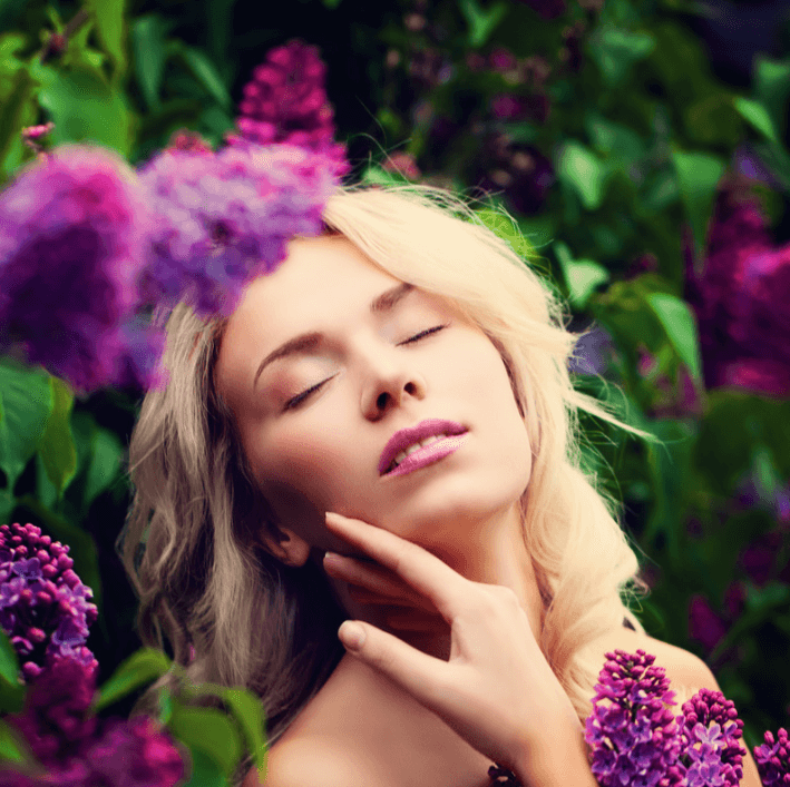 Vrouw met een stralende huid omringd door paarse bloemen, die elegance en schoonheid uitstraalt.