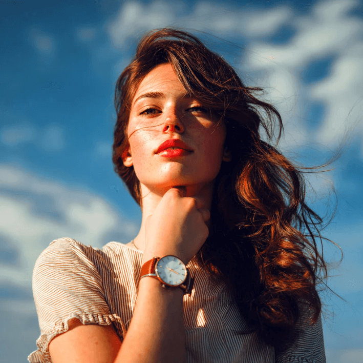 Vrouw met lang haar in een zonnige omgeving, met een premium volumizing lippenstift en een elegante horloge.
