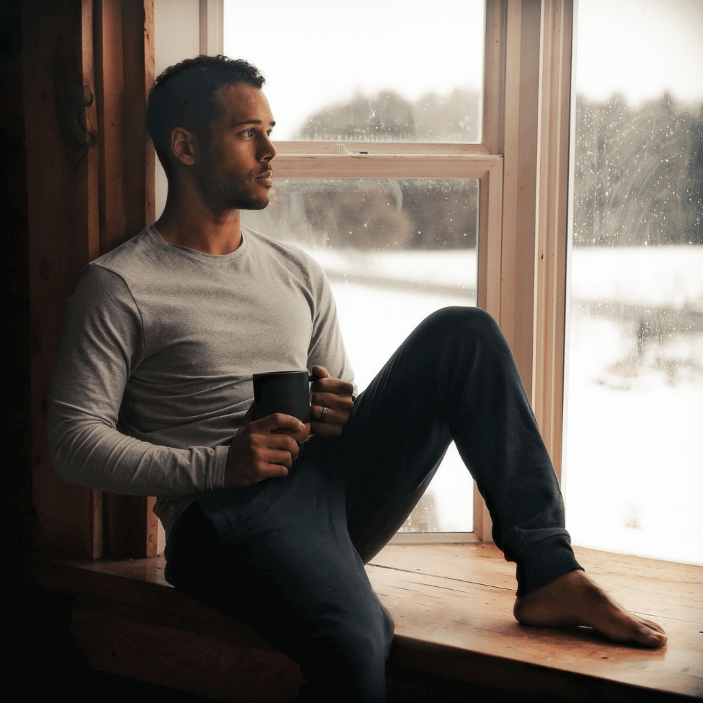 Man in a cozy sweater holding a mug, sitting by the window with a serene winter view.