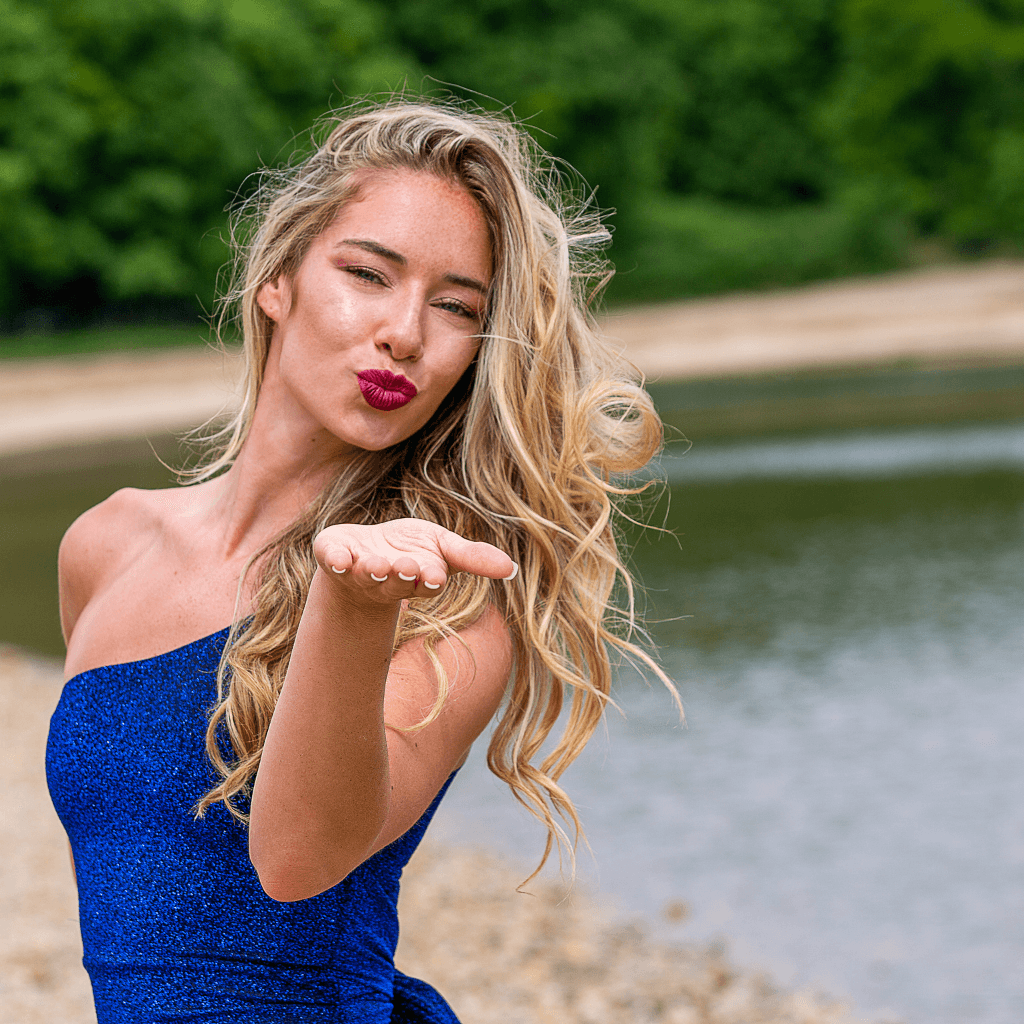 Vrouw in een blauwe jurk die een kus gooit bij een waterpartij in een natuurlijke omgeving.