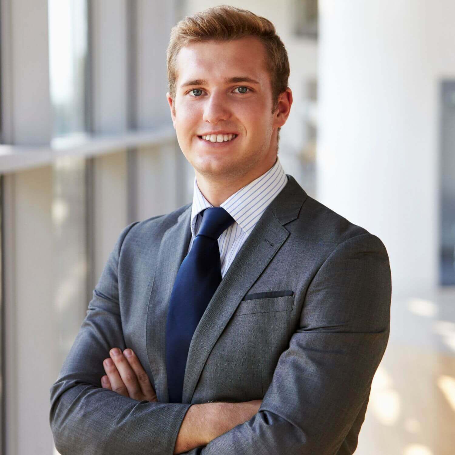 Professionele man in een grijs pak, glimlachend en met armen over elkaar, staand in een moderne kantooromgeving.